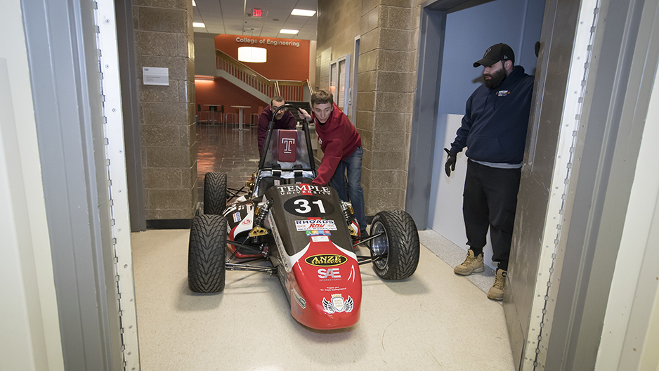 Two students pushing the race car past an elevator door. 