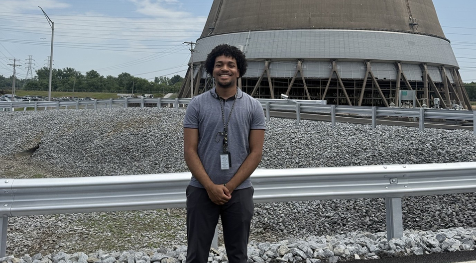 A photo of Adam Lawrence facing the camera in a gray polo and black pants. He is outdoors in front of a cooling tower.