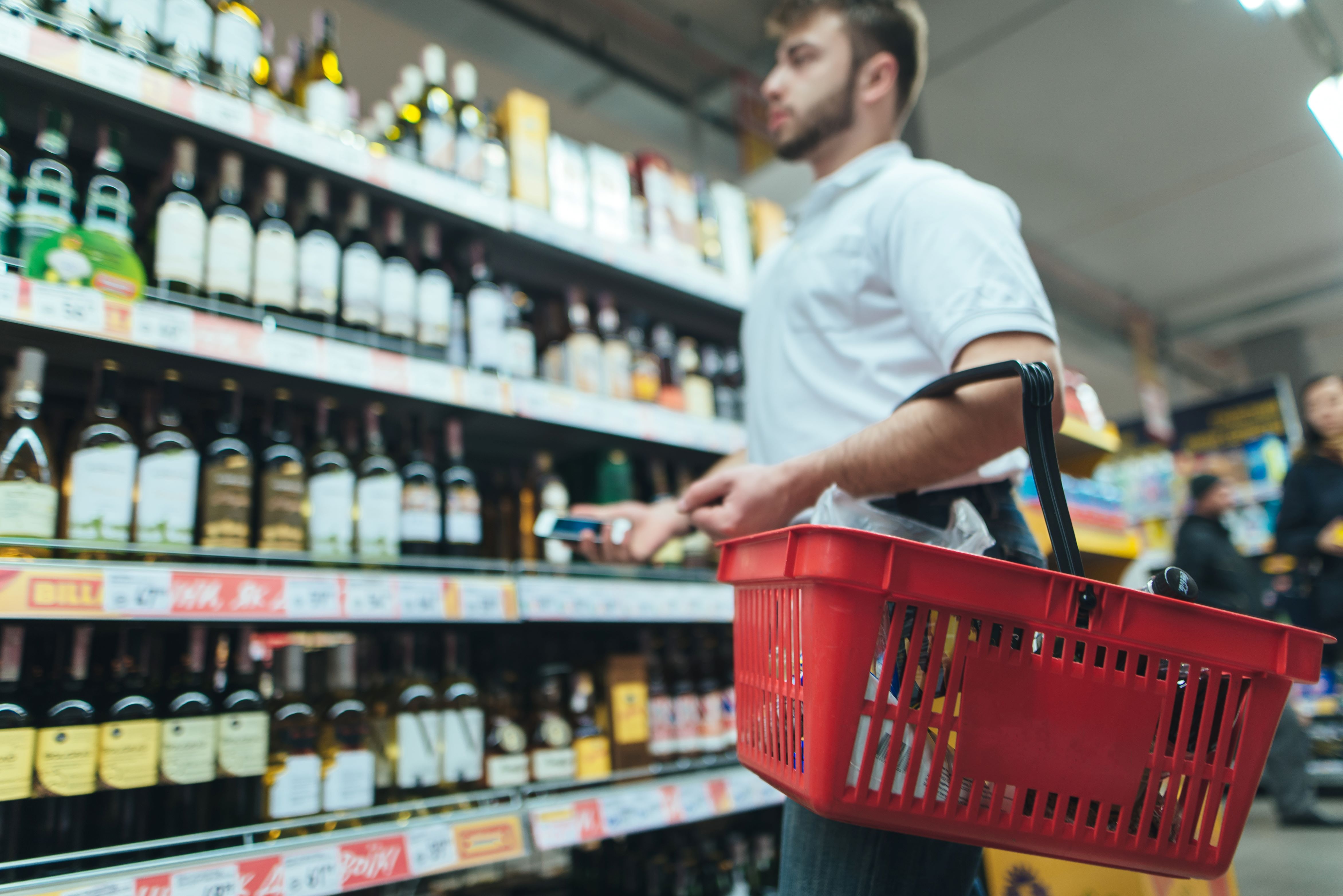 Man holds a red shopping basket in front of shelves of alchohol