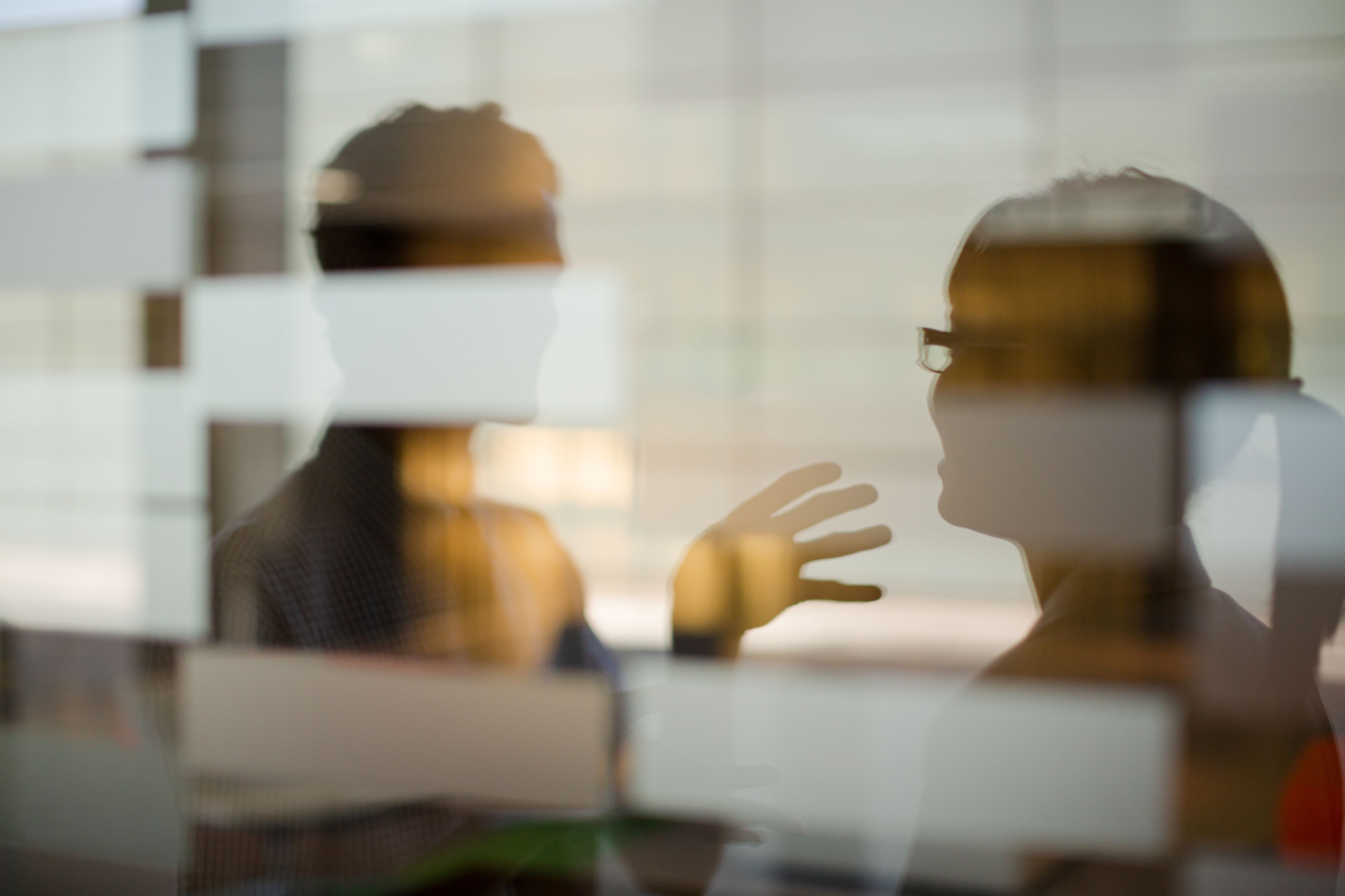 Two people with their faces obscured, speaking to each other behind a glass wall