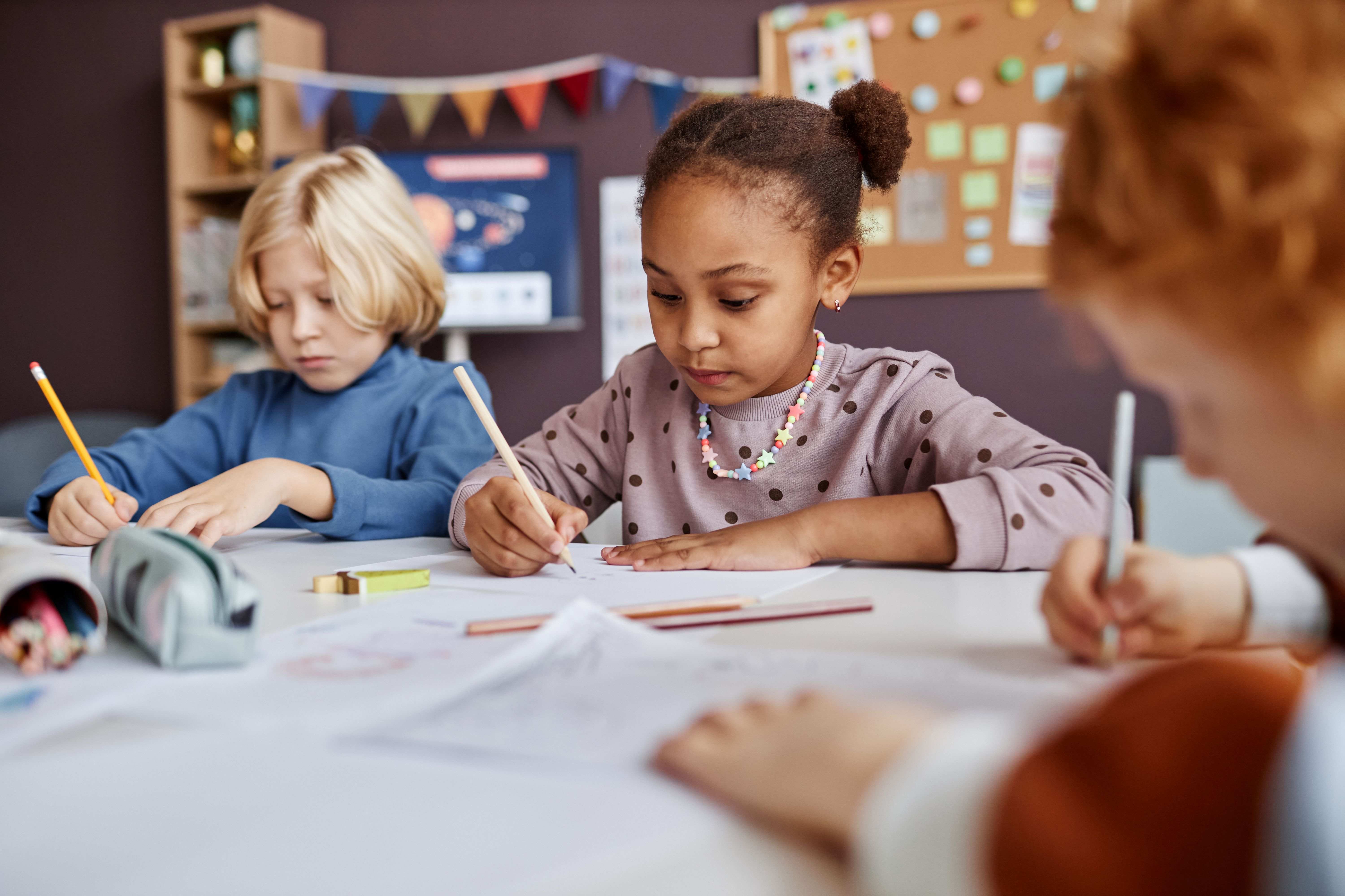 Three you children sitting at a table writing with pencils