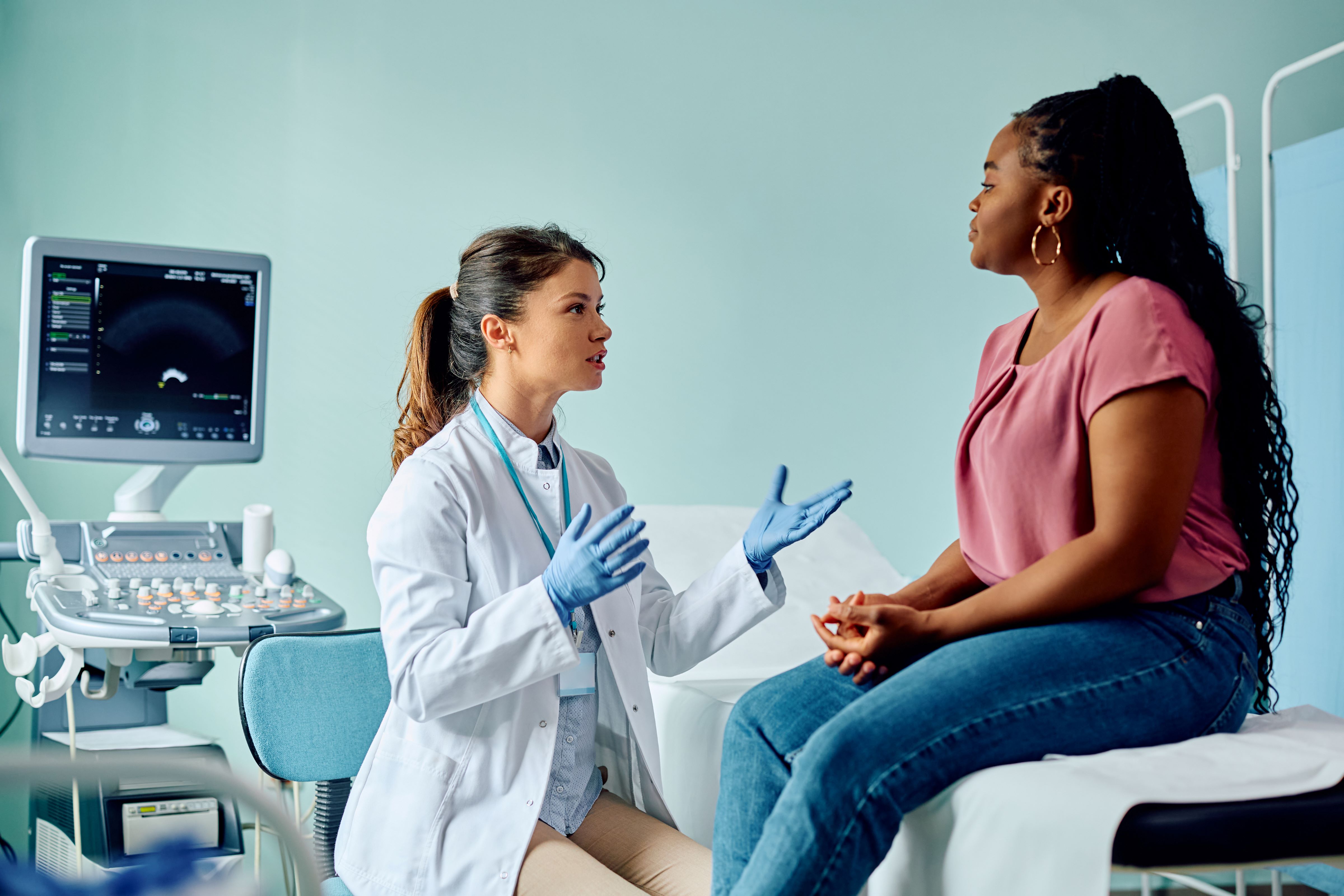 A woman with lighter skin and long brown hair in a ponytail wearing a white lab coat and blue gloves speaks to a black woman sitting on an exam table in a medical setting