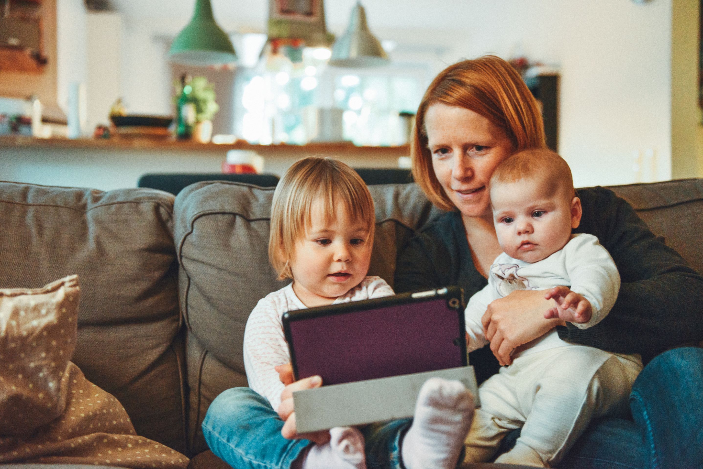 Woman with red hair sits with two children on her lap, a toddler and a baby, looking at a tablet