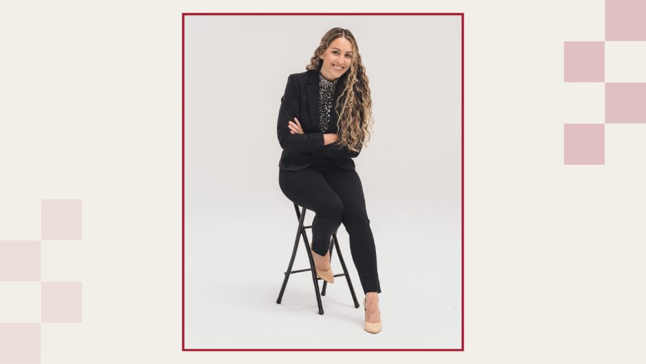 Amberly Caballero in a dark suit poses leaning forward on a high chair