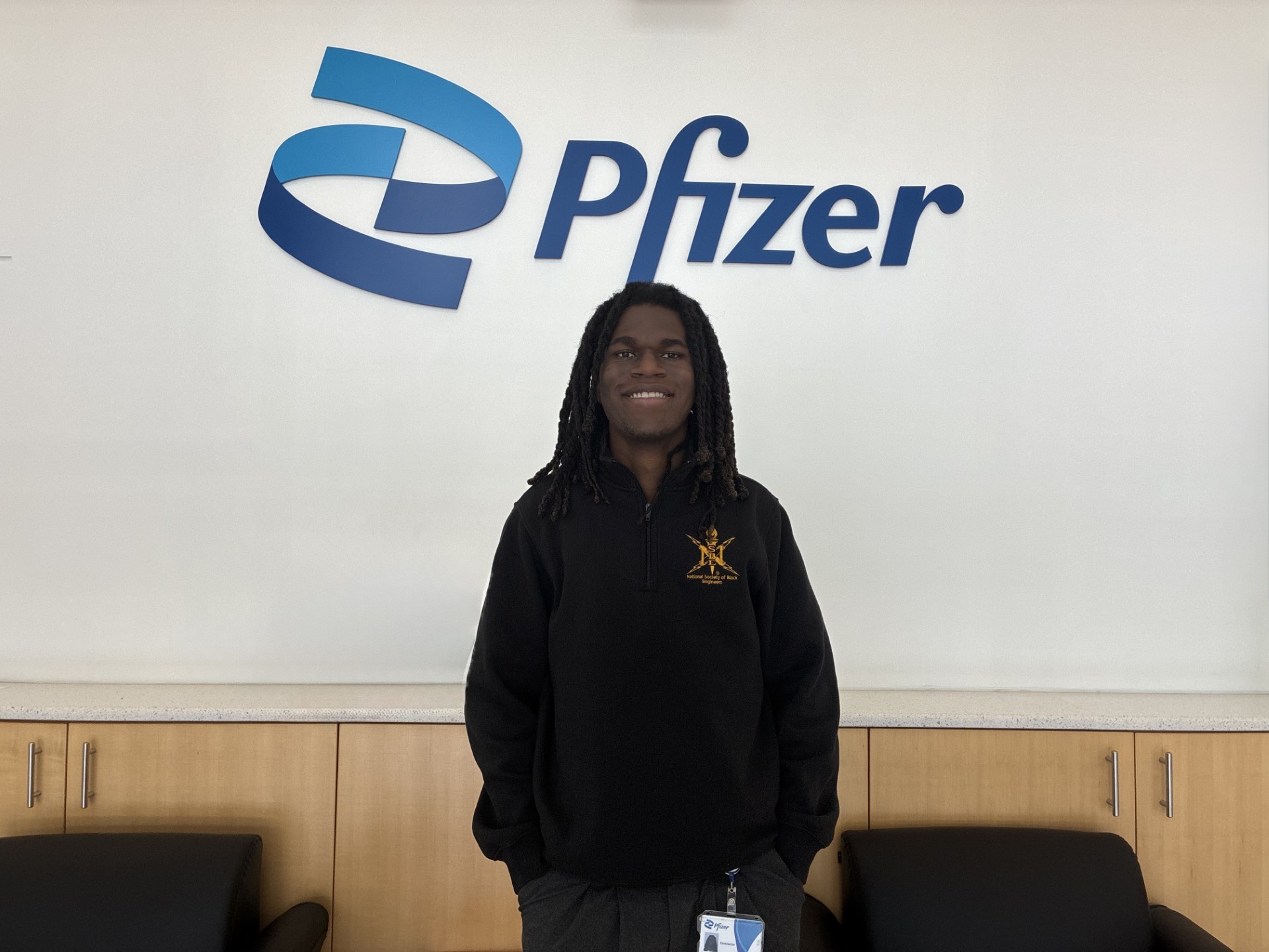 Brandon Henry standing in front of a white wall with a blue Pfizer logo on it. He is wearing a black quarter zip and smiling at the camera.