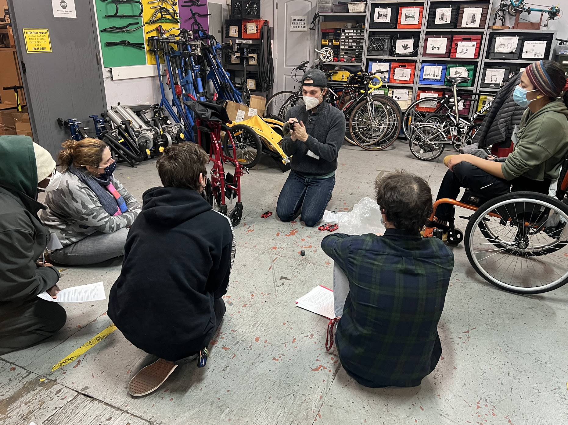 Six people at a wheelchair repair workshop. The instructor is repairing a manual wheelchair. One of the students is using a wheelchair.