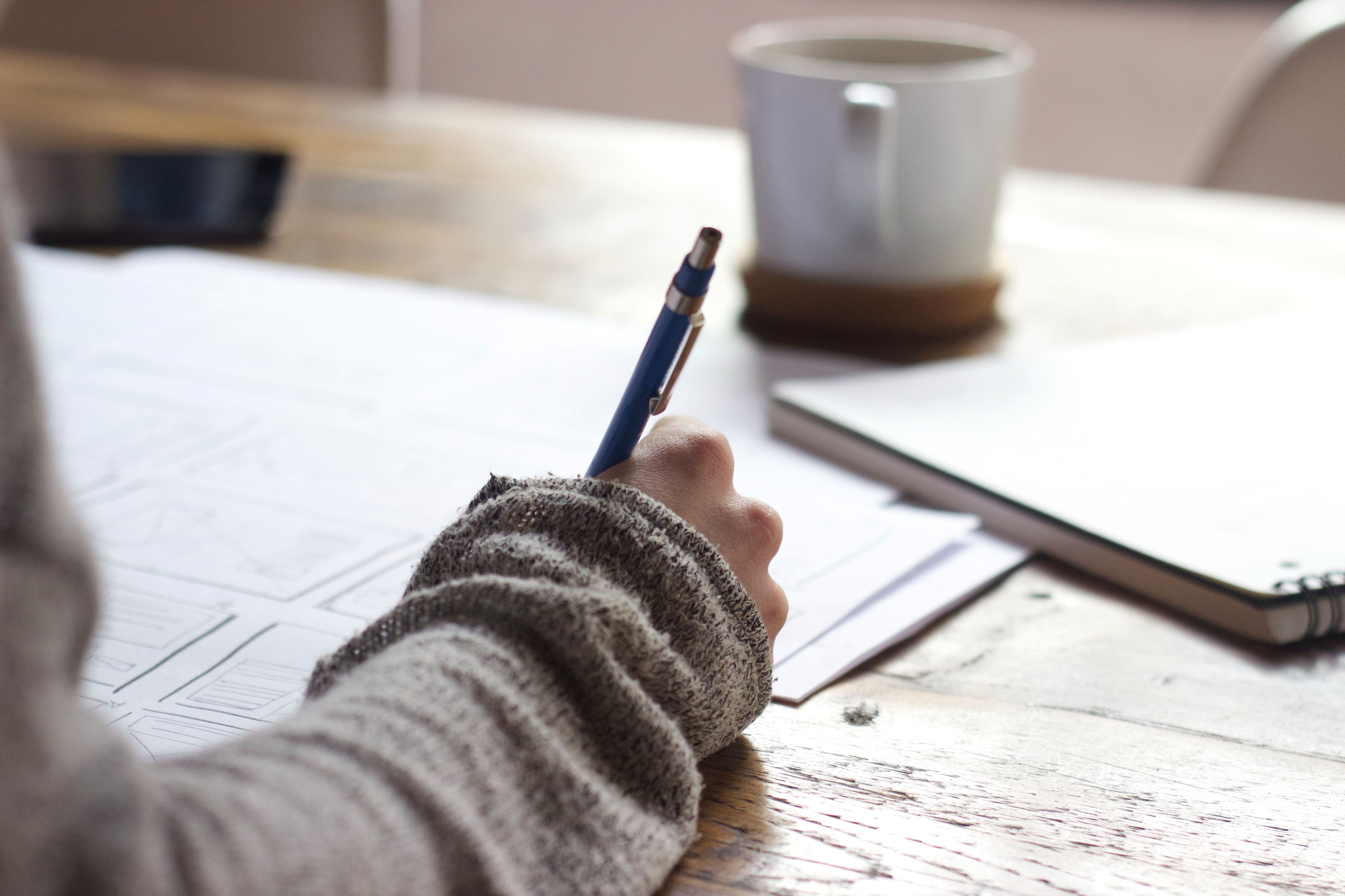 A hand holds a pen to paper, with a mug in the background