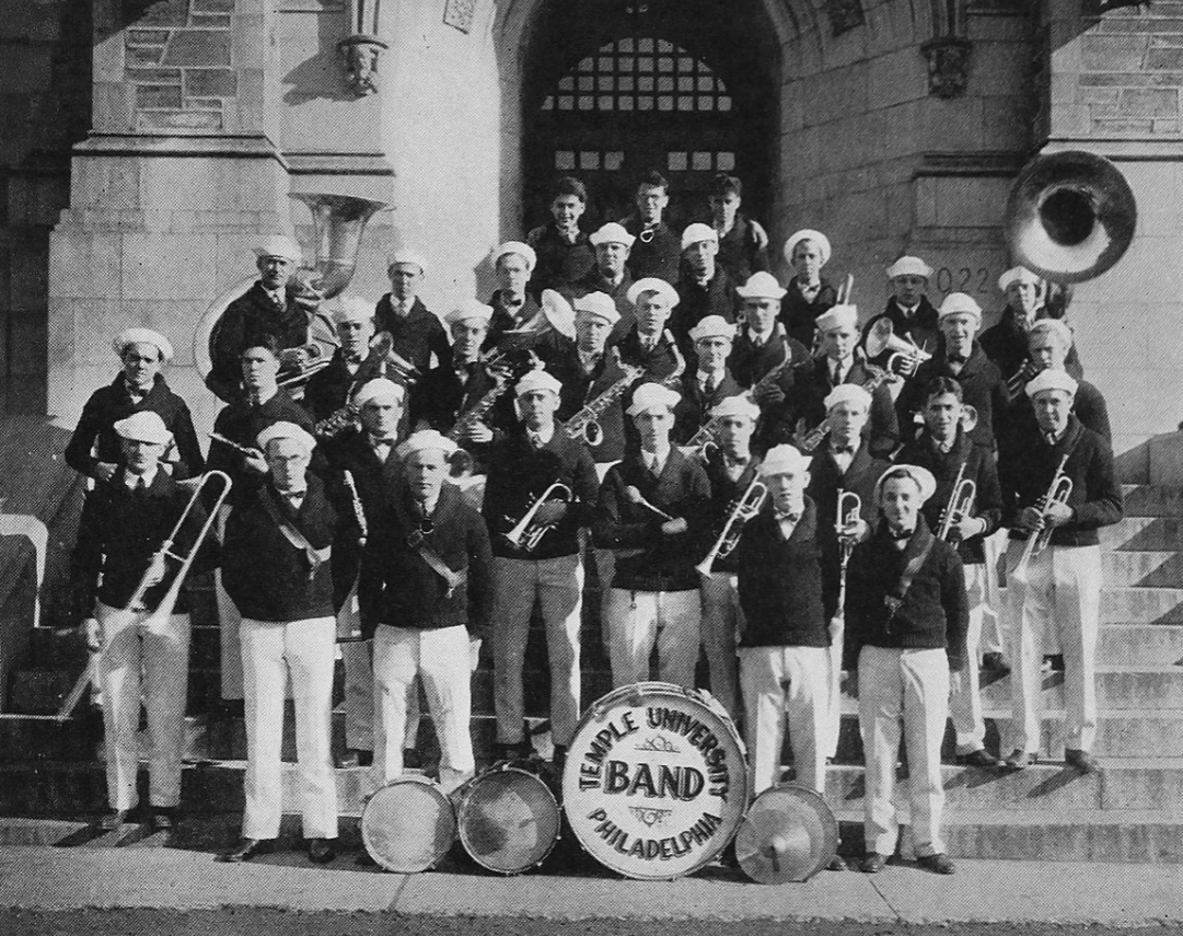 A group of young men standing on the steps of Sullivan Hall, posing with their instruments behind a bass drum which reads "Temple University Band Philadelphia"