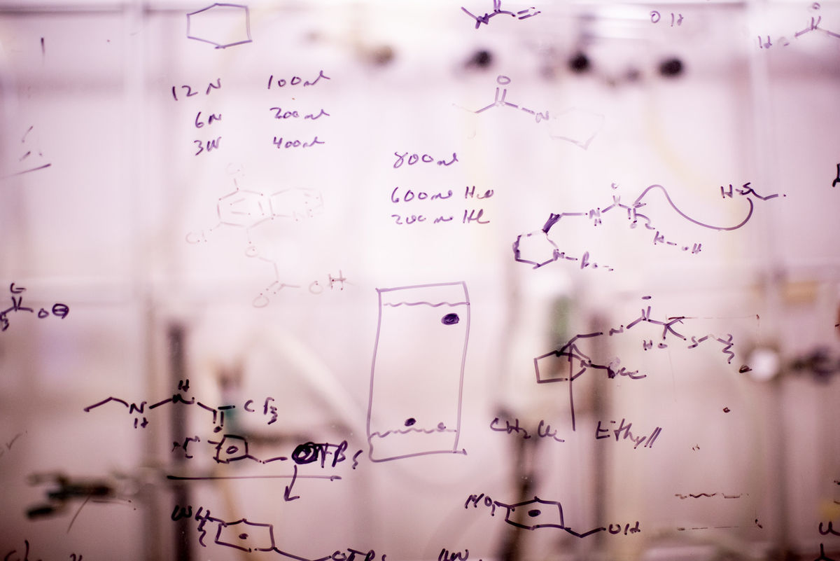 A semi-transparent piece of glass in front of a laboratory with dry erased scientific drawings on it.