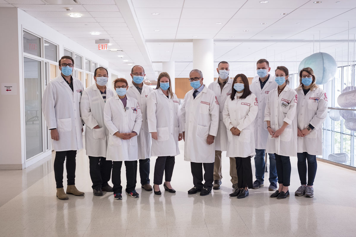 A group of scientists in white coats and masks stand shoulder-to-shoulder.