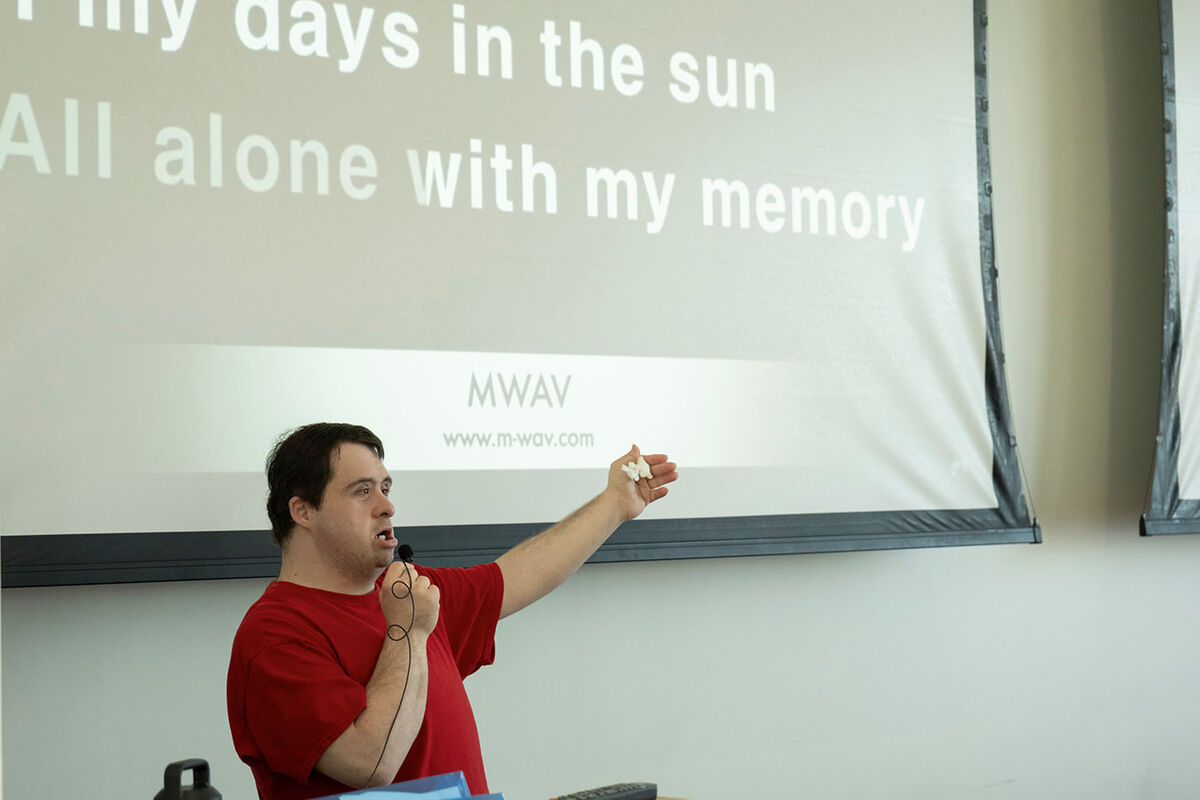 RJ, wearing a red t-shirt, sings into a hand-held microphone with one arm extended, at the front of an indoor space, next to a projected slide that reads, 