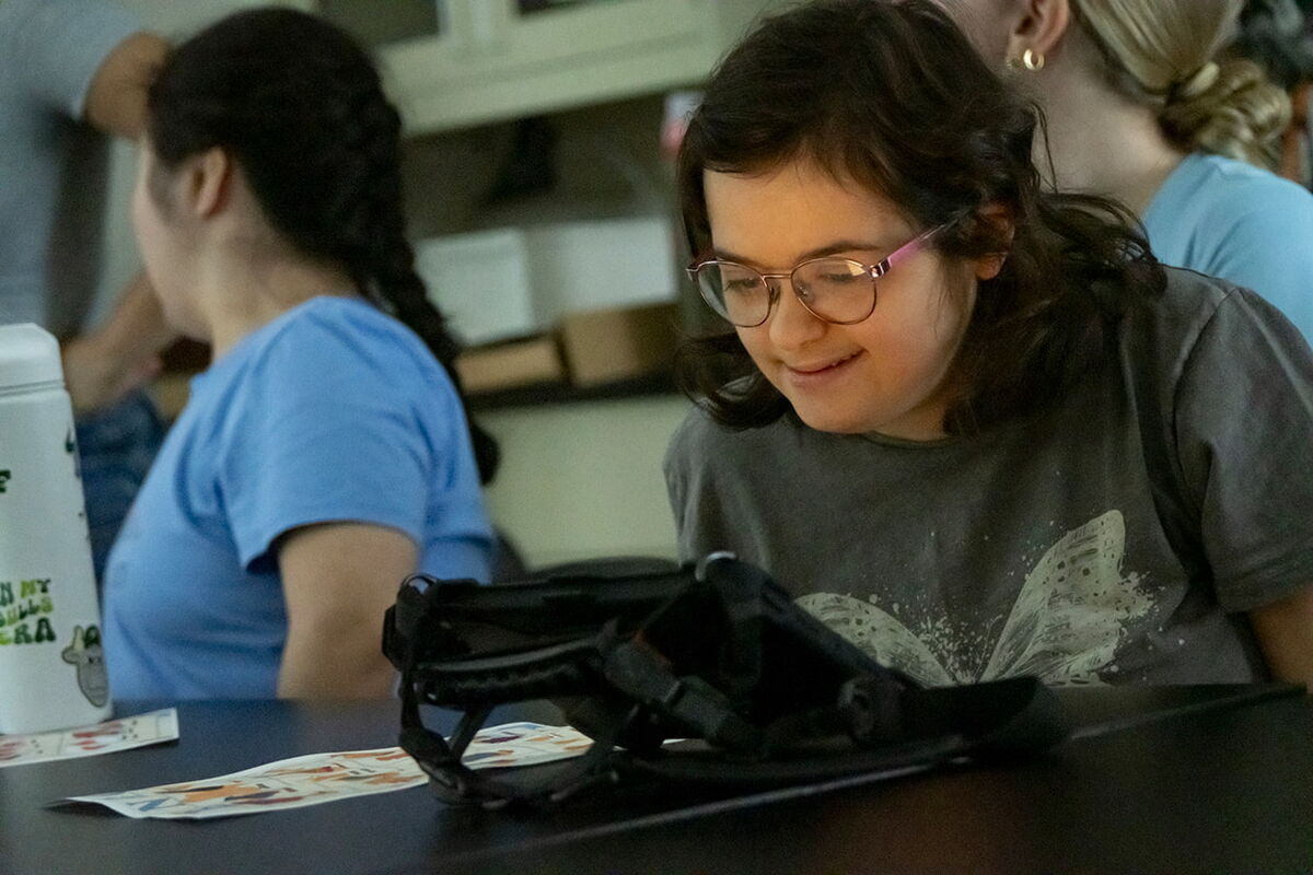 Ruby uses her communication device at her desk in a classtoom, a slight smile on her face. 