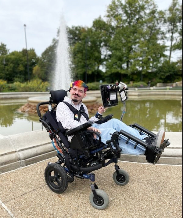 Mark Steidl, in a dress shirt and sporting beard, mustache and rainbow-colored hair, smiles in front of an outdoor fountain while using wheelchair with attached AAC device.