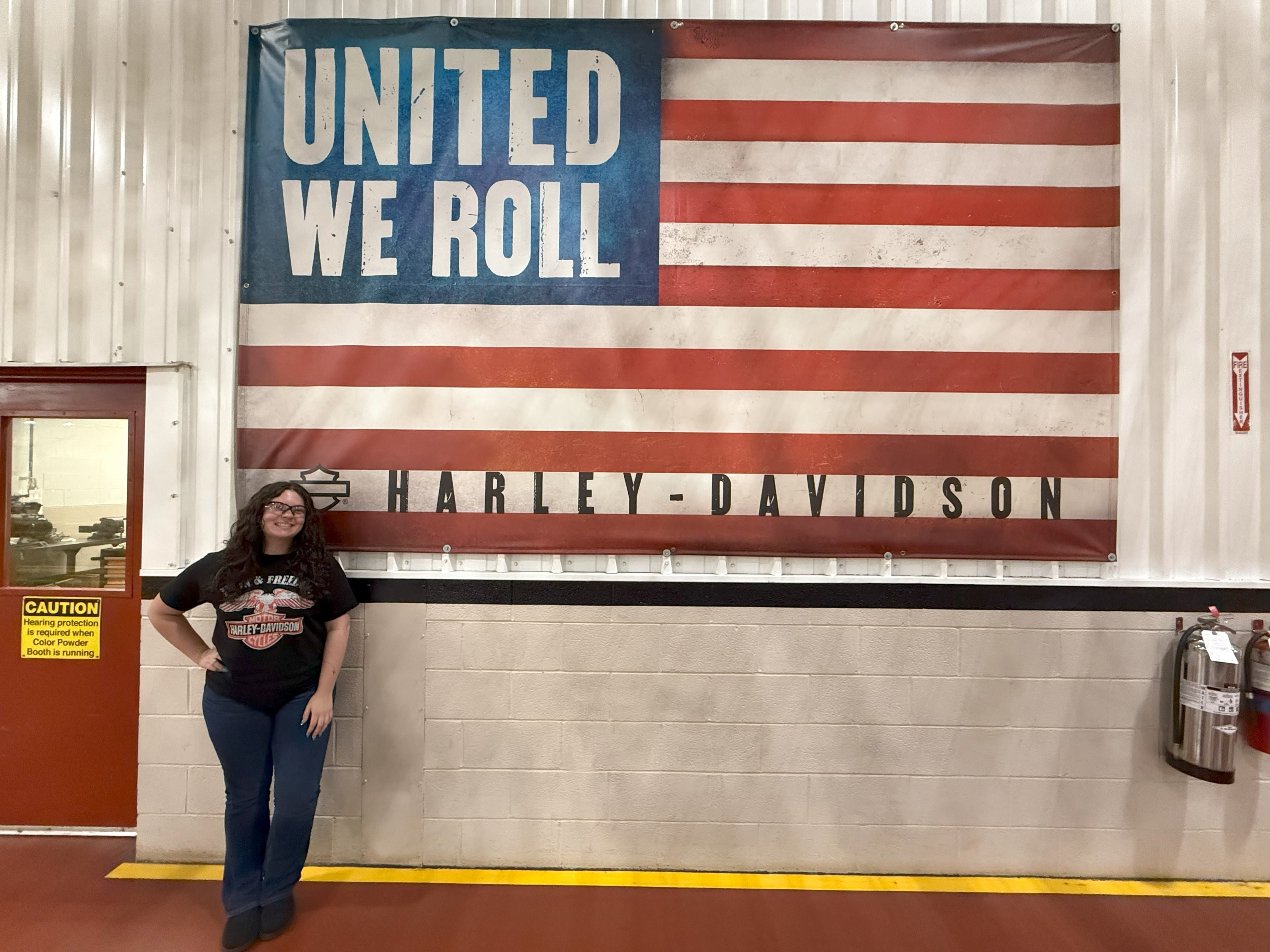 Brasten stands near an American flag that reads "United We Roll" and "Harley-Davidson."