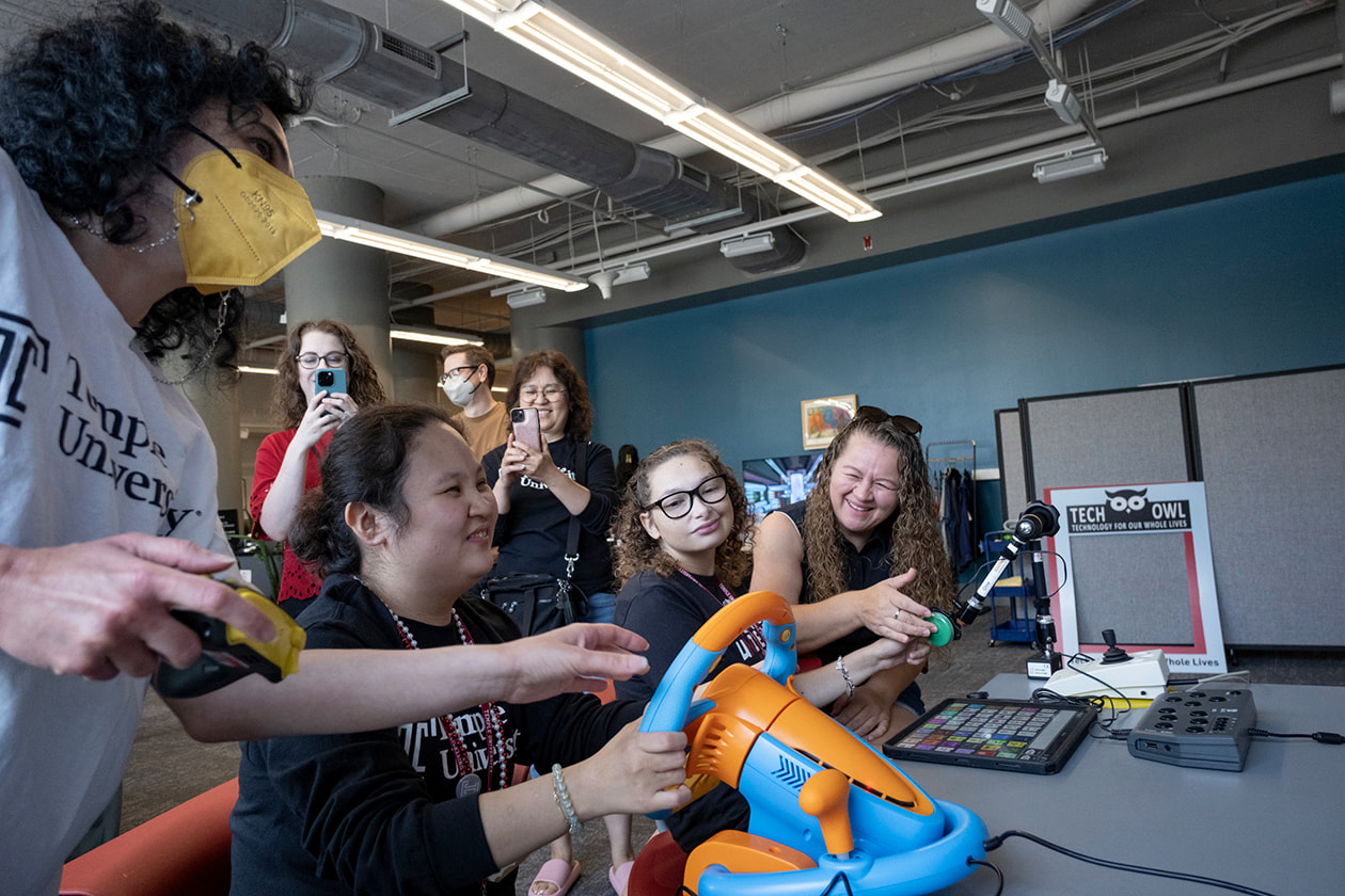 Elizabeth, left, and Alyssa, right, use adapted gaming devices in the TechOWL Community Space. A staff member assists Alyssa. Behind them, a staff member and caregiver take photos.