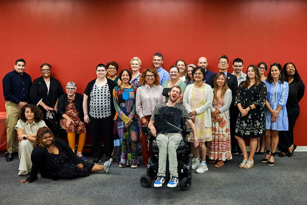 C2P2 graduates, former graduates, and IOD staff pose, smiling, in front of a red wall for a post-graduation photo. One graduate, Liam Holland, uses his wheelchair in the first row.