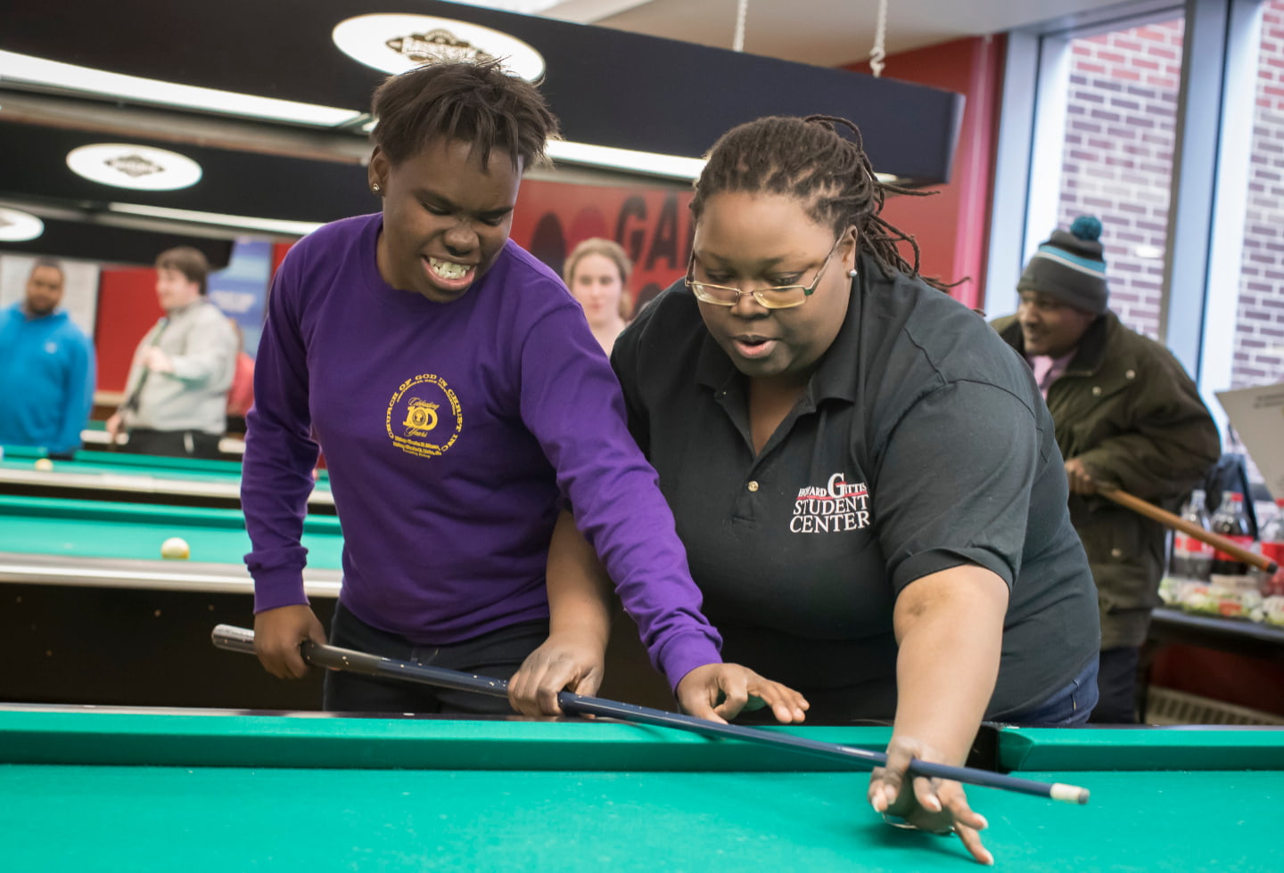 A student plays pool in the Howard Gittis Student Center gameroom, aided by a staff member who helps the student to grip the cue stick. 