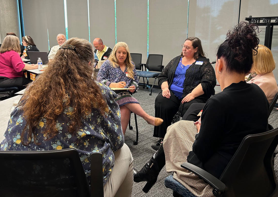 Four professionals in a conference room converse, with their chairs arranged in a circle
