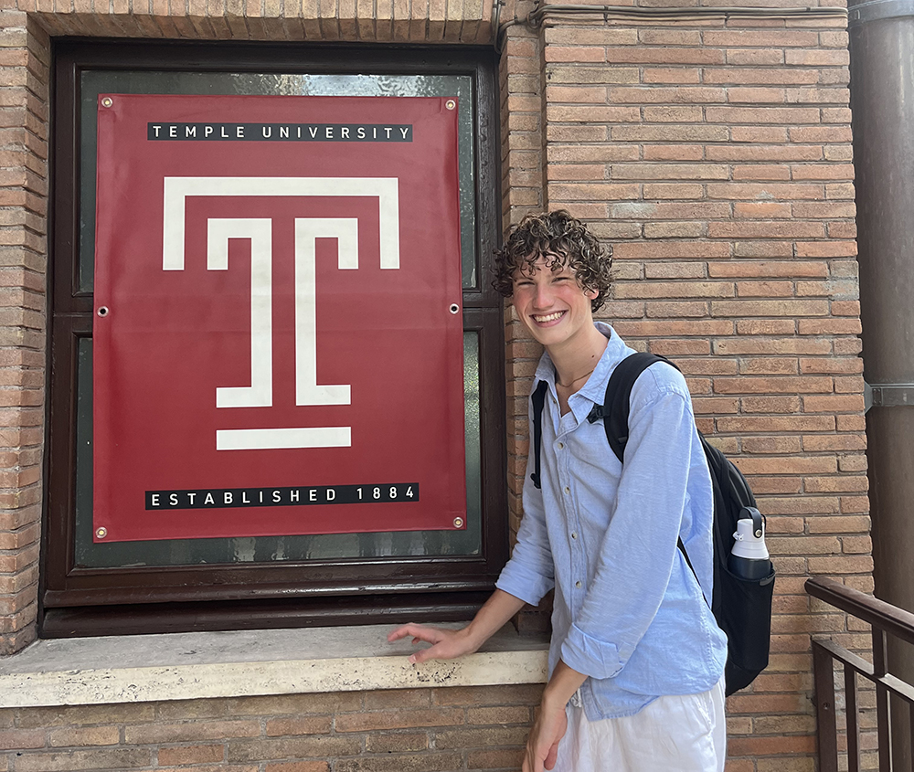 A student, Justin, is wearing a blue button down shirt with a black backpack. He is standing to the right of a Temple University sign on a building at Temples Rome campus.