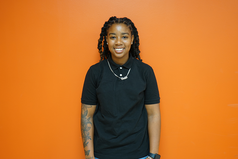 A headshot of Kayla Garrison. She is standing in front of an orange wall wearing a black polo shirt.