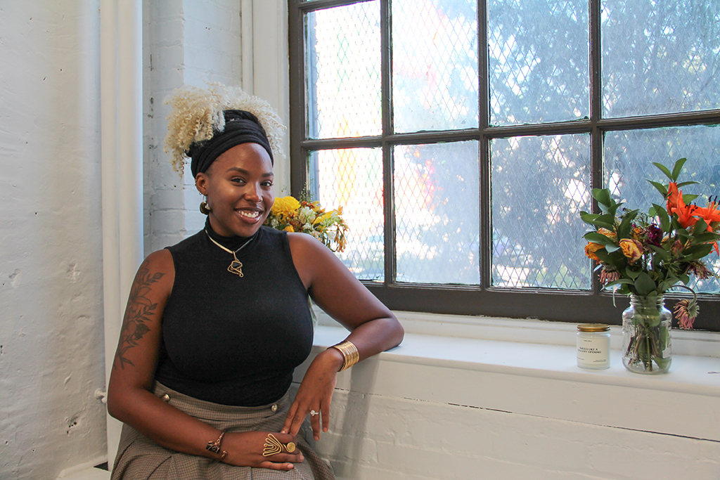 Chelsey Luster smiles at the camera. She is in Luster Gallery + Studio, a white-walled exhibition space. She sits next to a window.