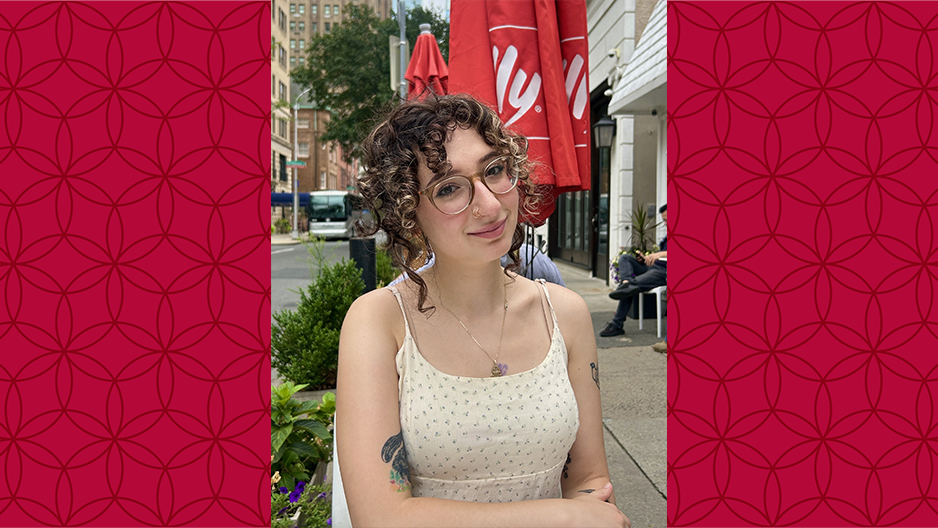 image of Maia sitting at a table on Temple campus wearing a cream dress and glasses smiling at the camera