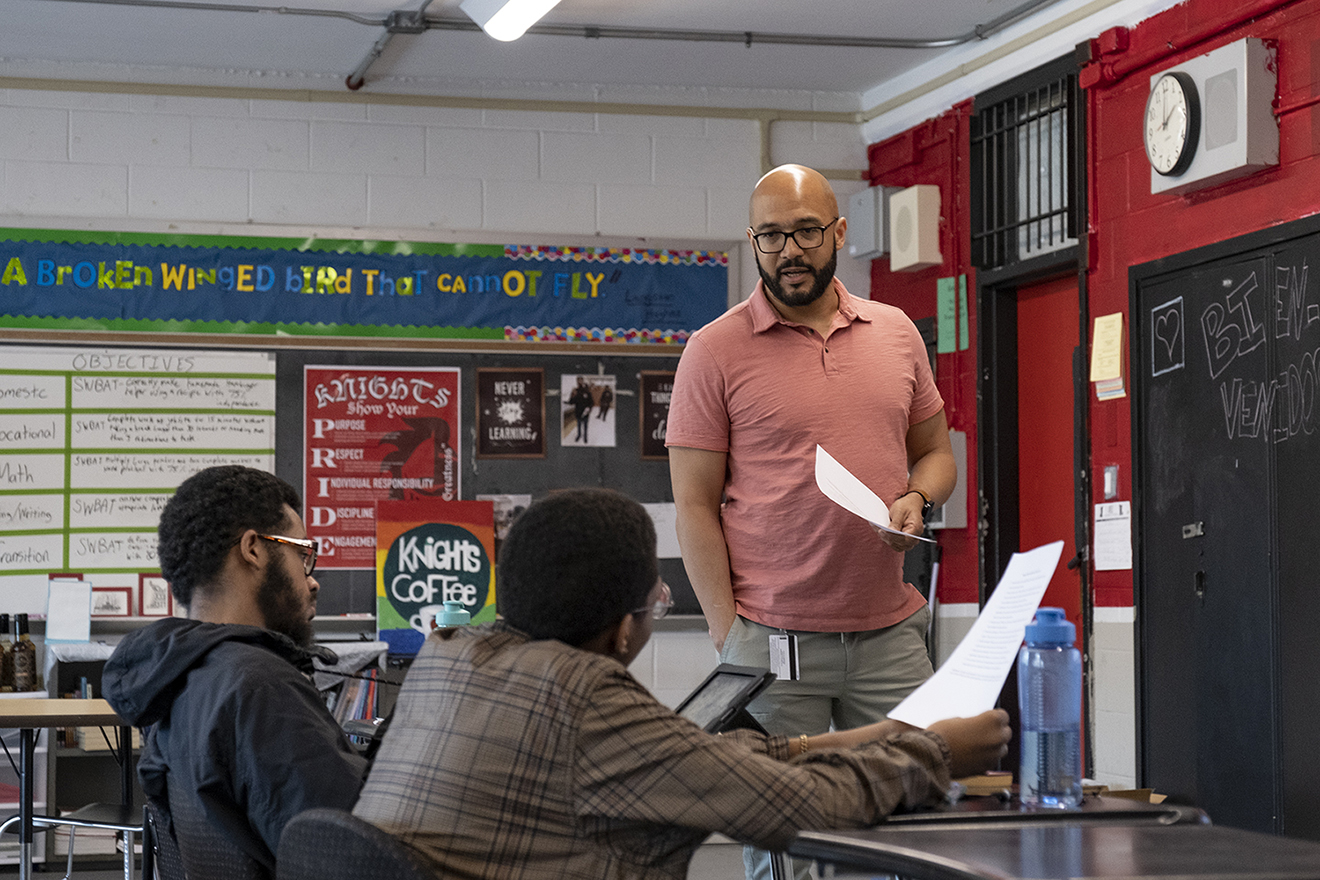 A man stands at the front of a classroom with teenaged students