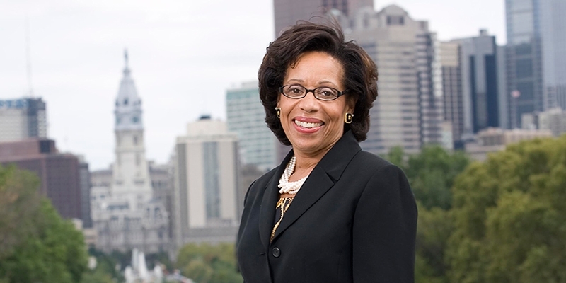 A woman in glasses and a black jacket standing before Philadelphia's skyline.
