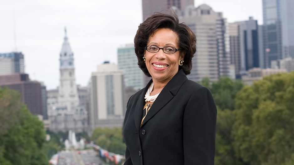 A woman in glasses and a black jacket standing before Philadelphia's skyline.