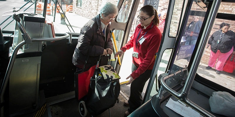 A young woman helping an older woman carrying a cart safely board a bus.