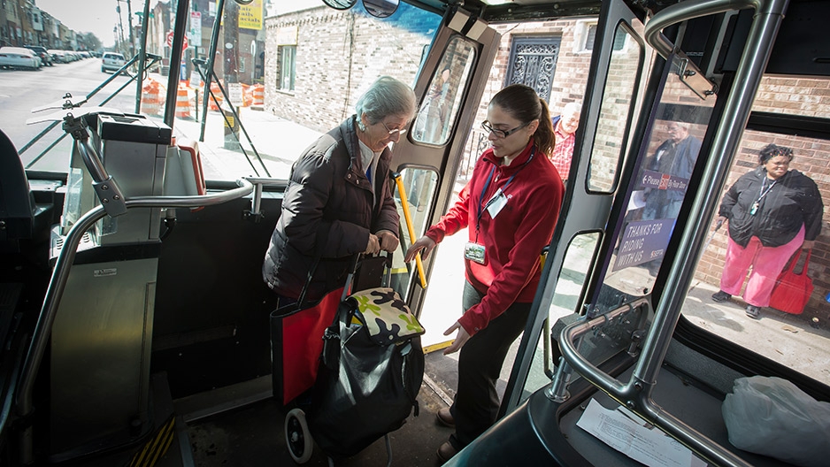 A young woman helping an older woman carrying a cart safely board a bus.