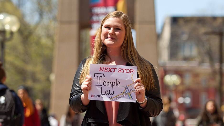 A woman in a black jacket holding a sign in front of Temple’s bell tower.