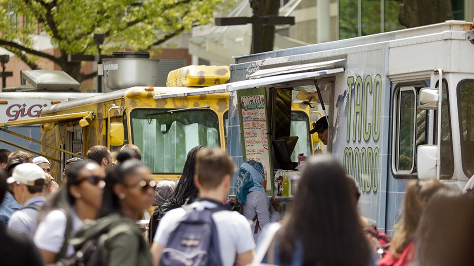 A crowd of people in front of a row of food trucks on campus. 