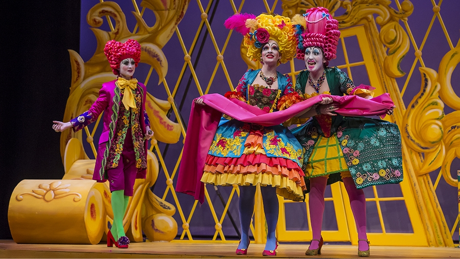Three opera singers wearing brightly colored costumes and wigs performing on stage.