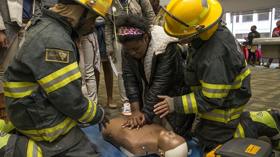 two firefighters teaching a female student how to perform CPR.