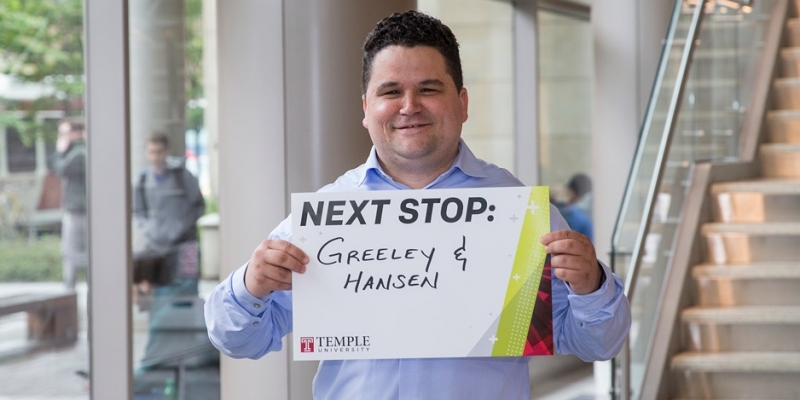 A man in a blue shirt holding a sign that reads Next Stop: Greeley and Hansen.