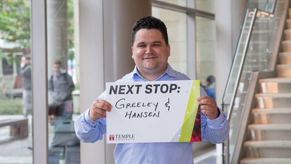 A man in a blue shirt holding a sign that reads Next Stop: Greeley and Hansen.