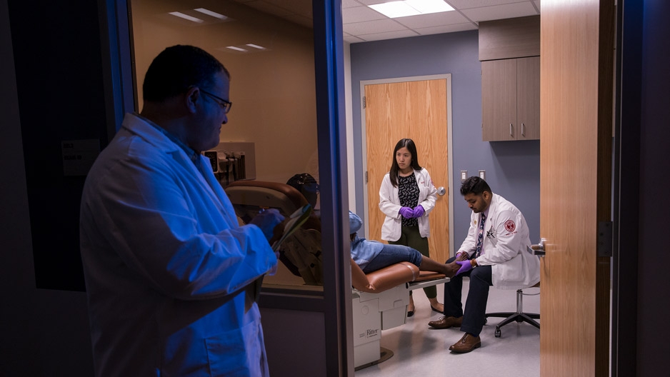 A physician watching two students work with a patient in a clinic room