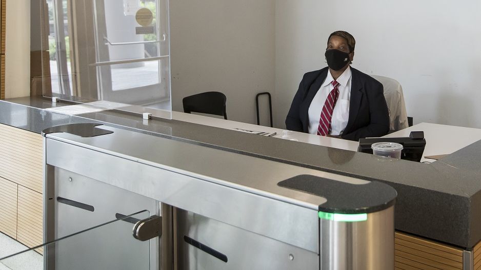 A security guard on campus wearing a mask and sitting behind a Plexiglas barrier at the entrance of a campus building. 