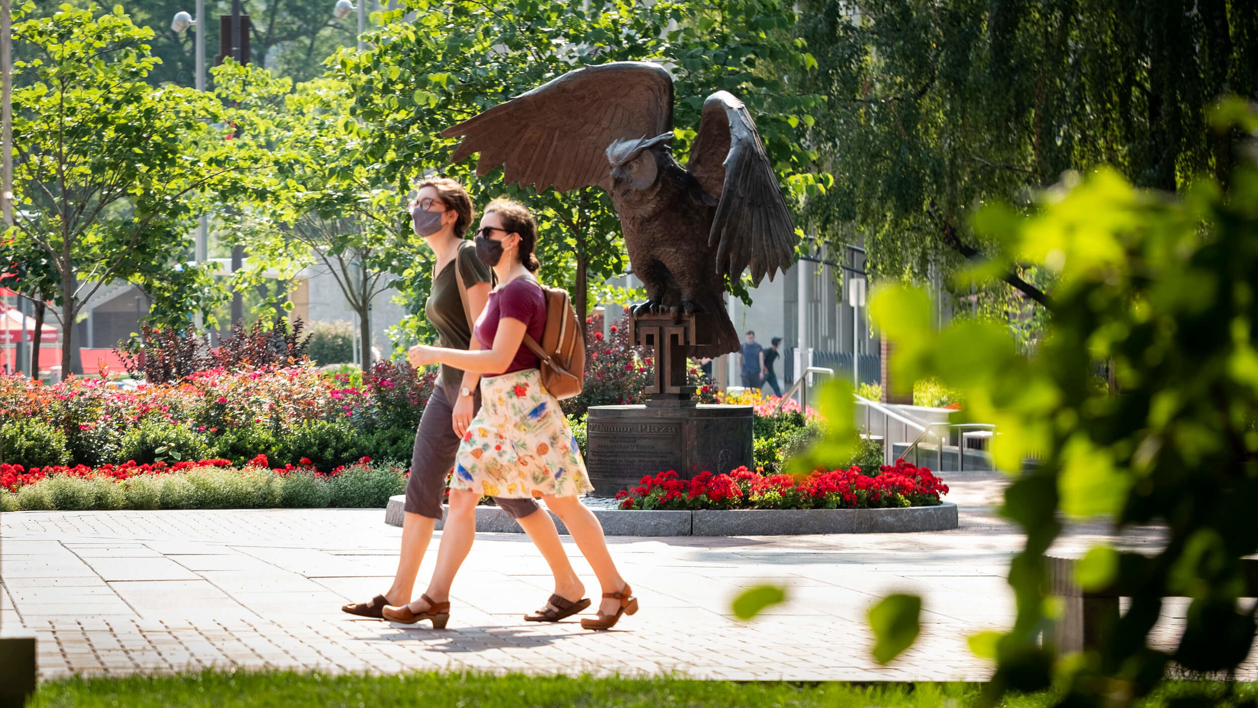 Two students wearing masks walk past an owl statue on Temple's Main Campus.