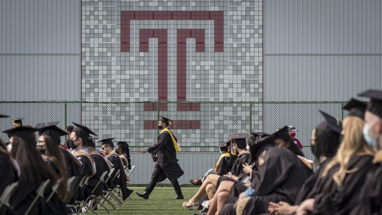 a graduate in a robe and cap walks in front of a large Temple T