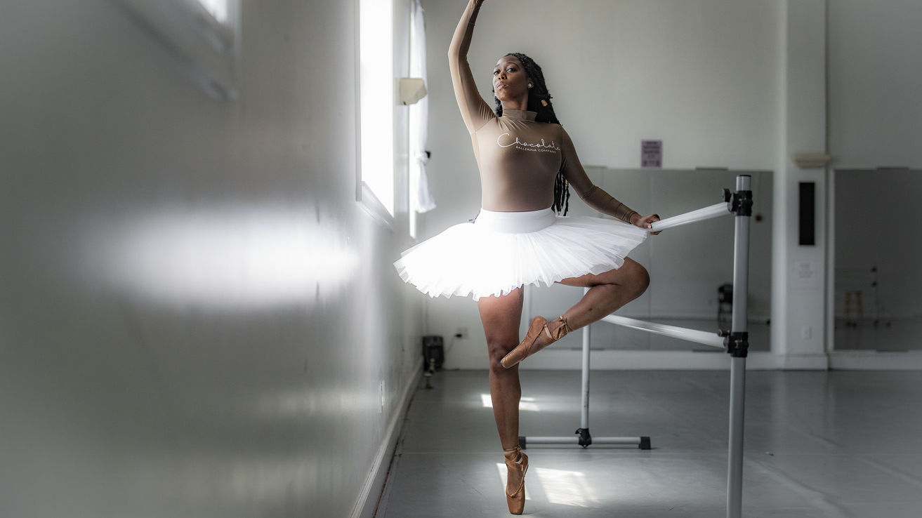 A Black woman in white tutu practicing ballet at a barre in a dance studio.