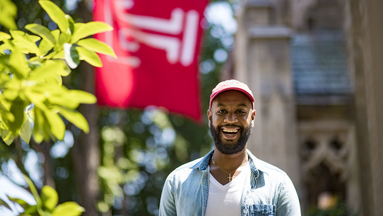 Brice Izyah smiling, wearing a red hat and standing under a Temple flag.