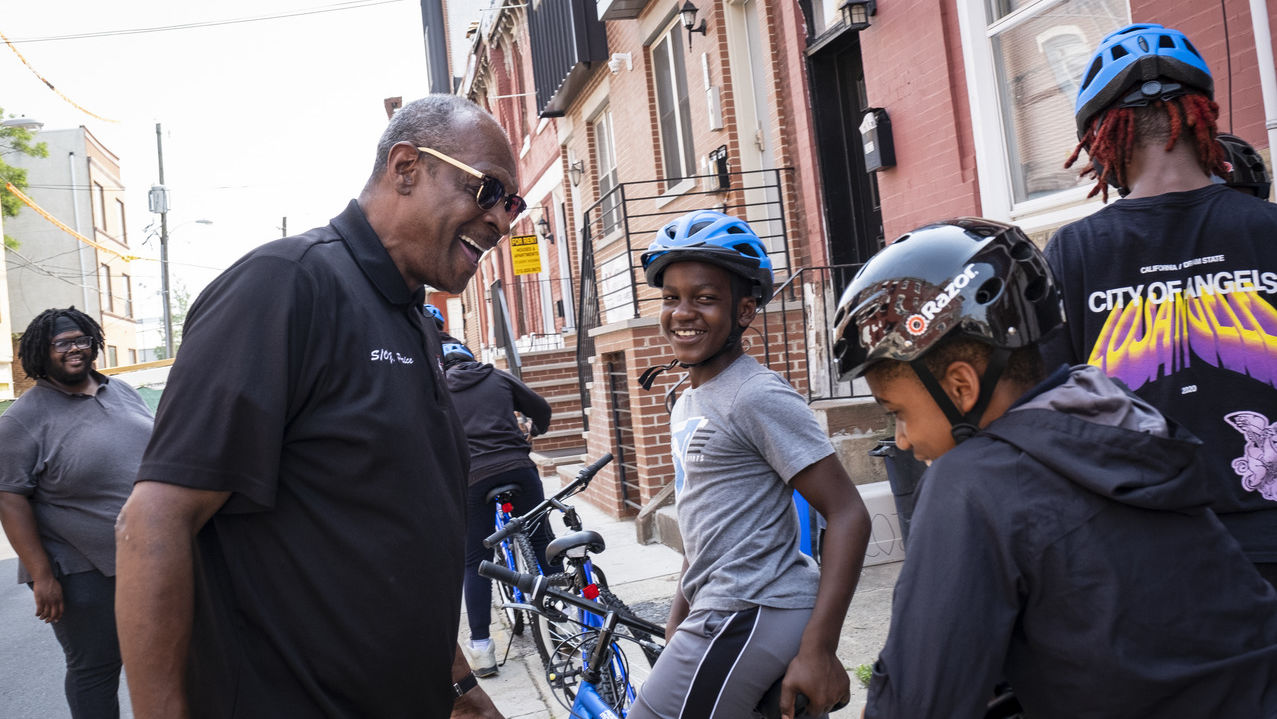 A smiling man in a black shirt with three happy children wearing helmets, sitting on bicycles during a Temple Urban Bike Team practice. 