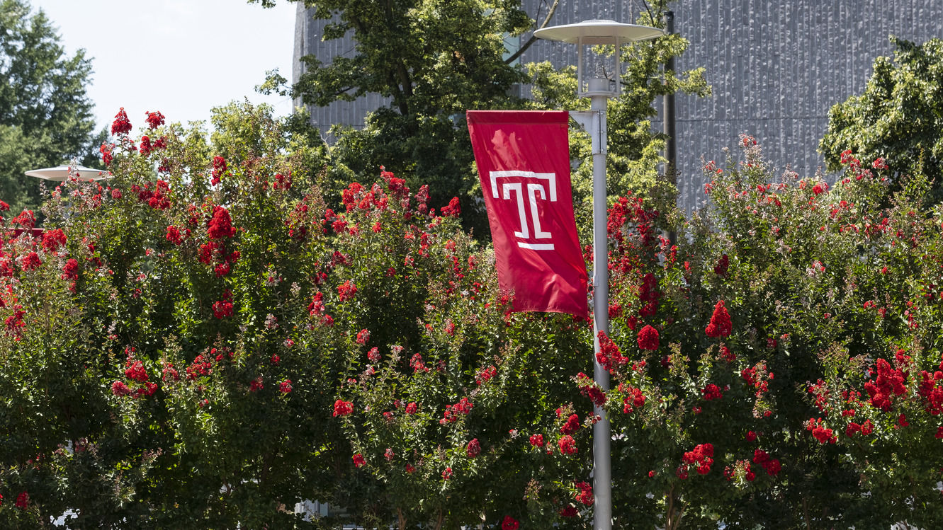 Summer on-campus stock photo pictured with a Temple flag and red flowers visible.