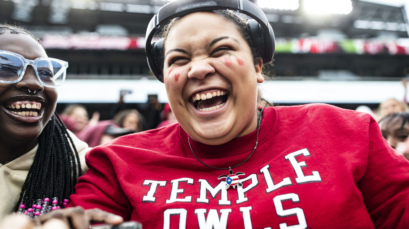 Image of a college student smiling, wearing a cherry and white shirt, seated at a Temple University homecoming event. 