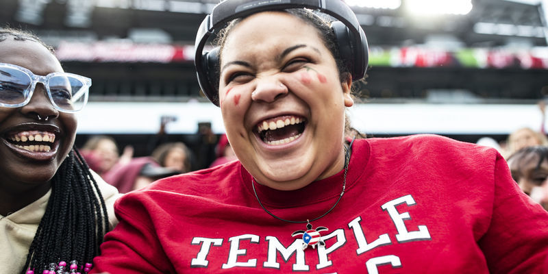 Image of a college student smiling, wearing a cherry and white shirt, seated at a Temple University homecoming event.