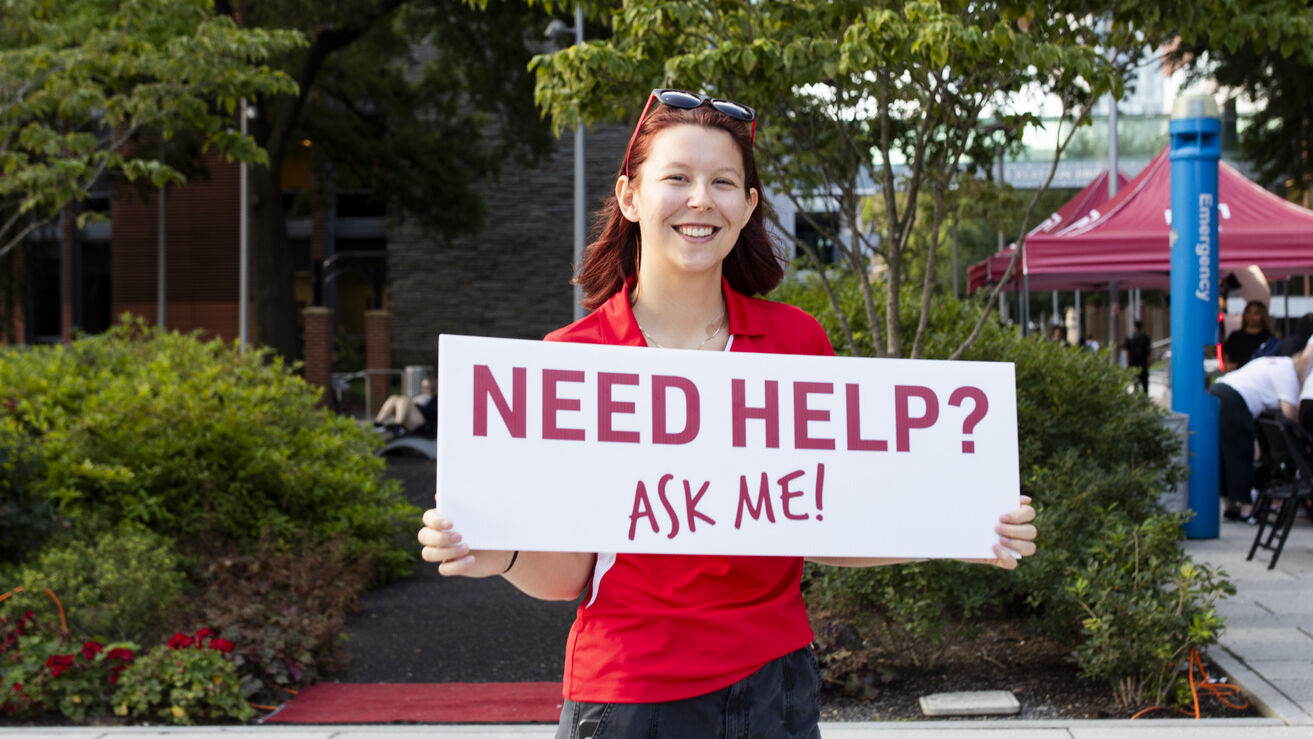 Student in a red polo holds a sign saying "Need help? Ask me!"