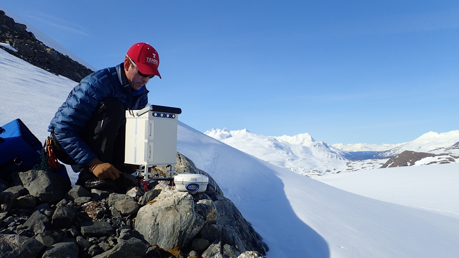 A man in a red Temple baseball cap perched on rocks with a snowy backdrop.