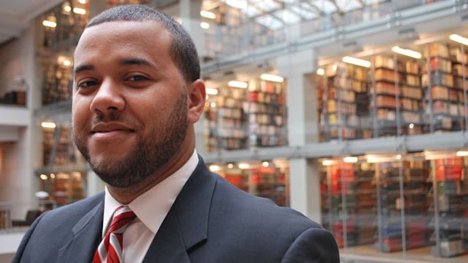 Nyron Crawford wearing a suit and tie with bookshelves in the background. 